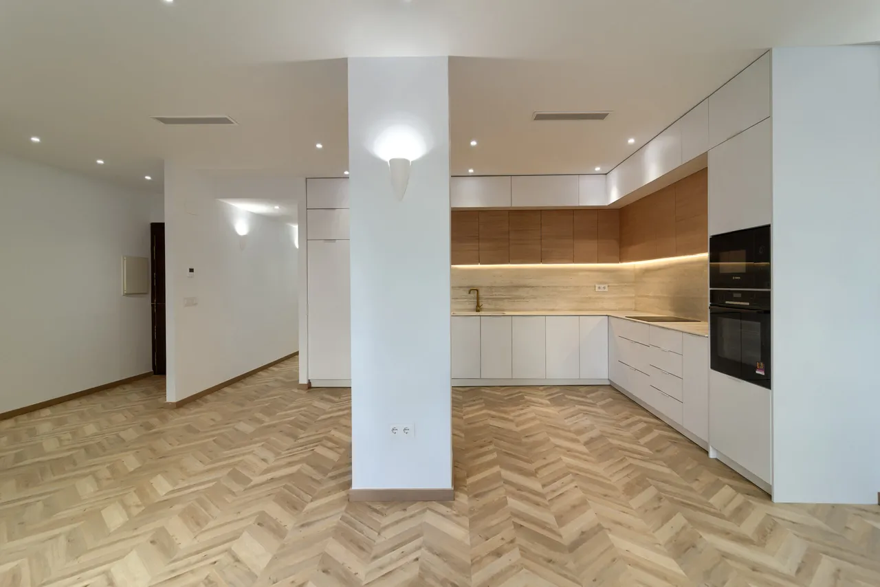 Cocina abierta con muebles blancos mate, frente de madera y suelo en espiga.