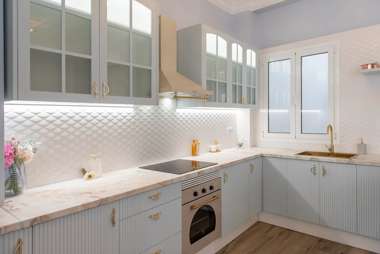 Cocina reformada con frentes estriados, puertas acristaladas, encimera efecto mármol y grifería en latón.