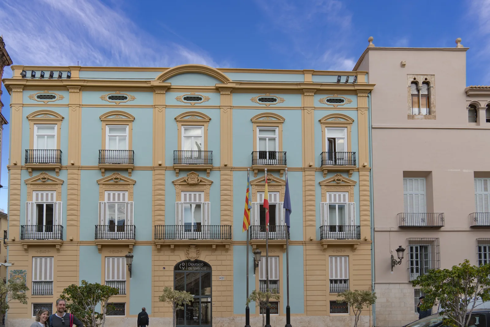 Fachada de un edificio de varias plantas en tonos azul claro y crema, con balcones de forja, ventanas de carpintería blanca, tres banderas sobre mástiles ante la puerta principal y varias personas en la acera.