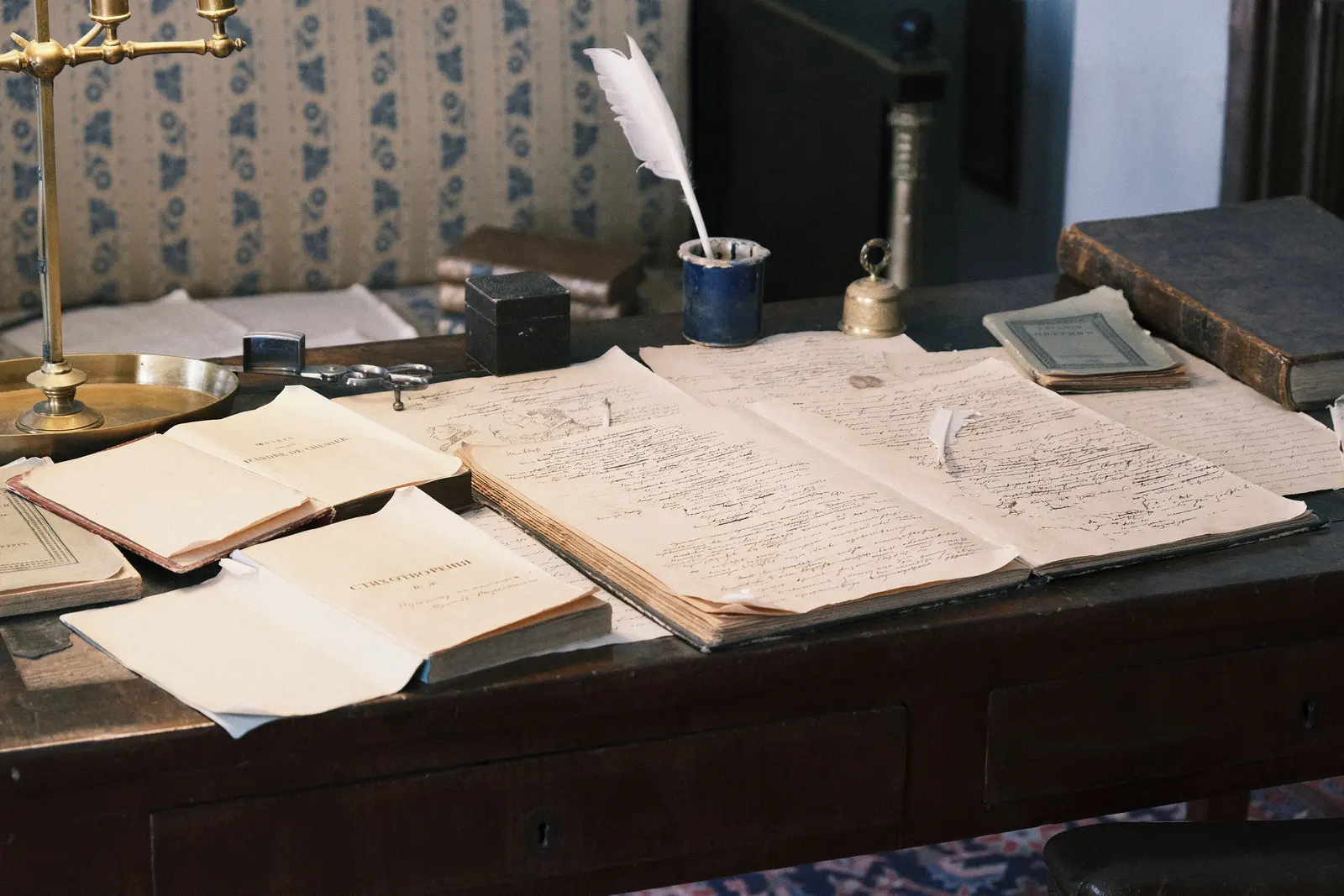 Mesa de madera oscura cubierta con documentos manuscritos antiguos extendidos, un tintero azul con pluma de ave blanca, una campanilla de latón, varios libros cerrados, unas tijeras y un candelabro de latón junto a una butaca tapizada.