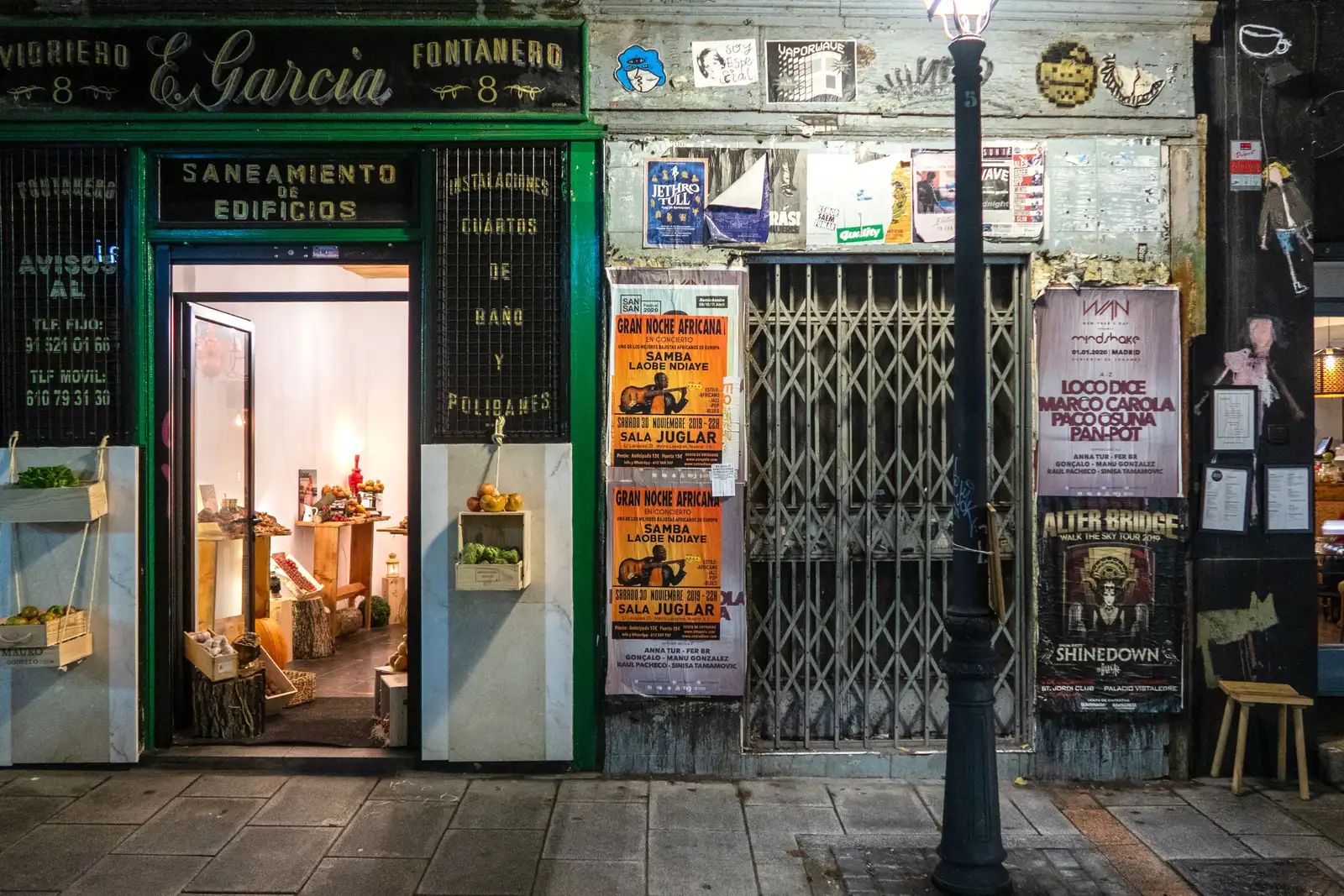Fachada de un local comercial a pie de calle, fotografiada de noche: a la izquierda, la entrada iluminada de una tienda con cajas de fruta y verdura junto a un rótulo; a la derecha, un hueco cerrado con rejilla y persiana metálica cubierto de carteles, y una farola de fundición en primer plano.