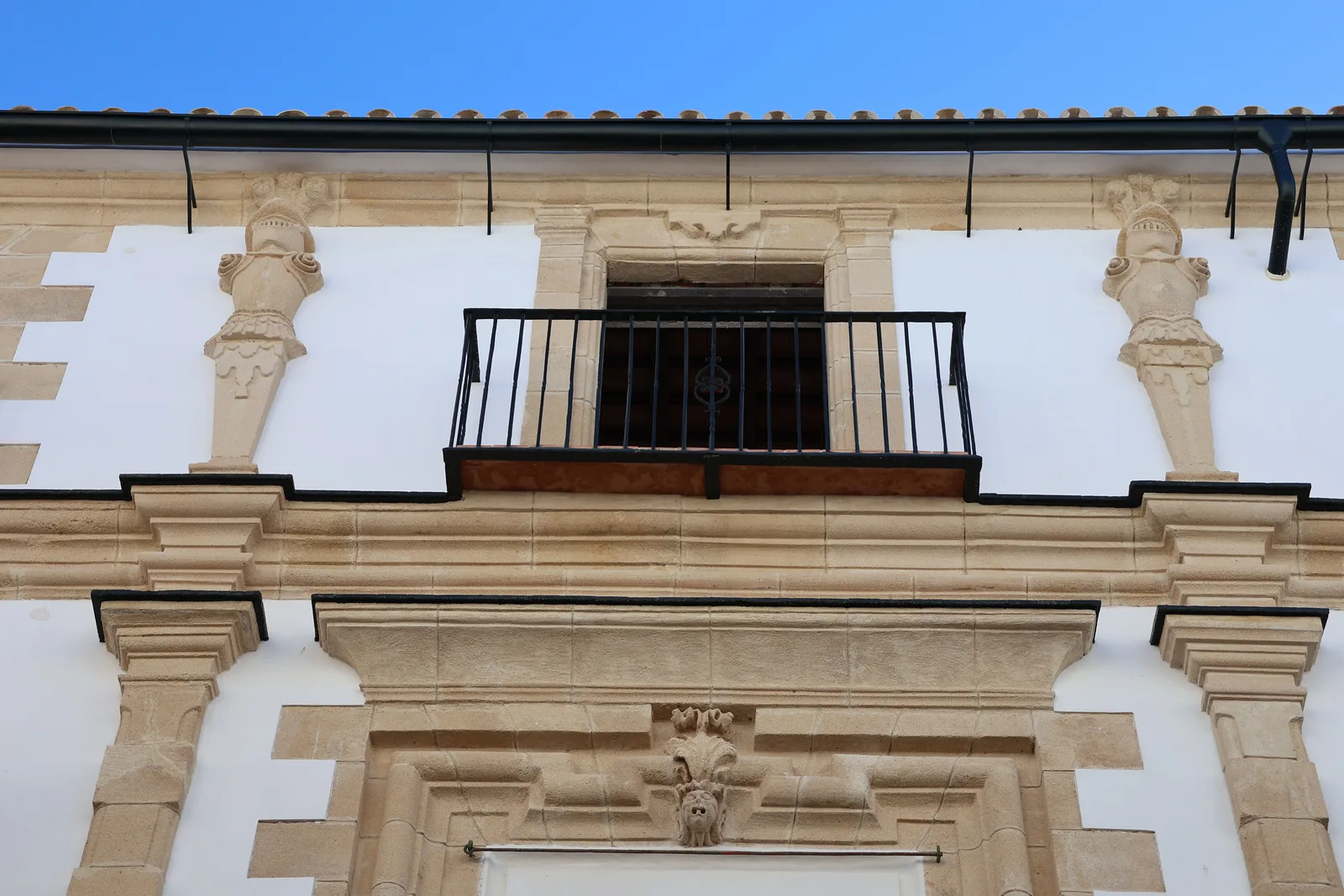 Vista frontal de la fachada de un edificio, con muro blanco y elementos de piedra, un balcón central con barandilla metálica y dos figuras ornamentales a sus lados, bajo un cielo azul despejado.