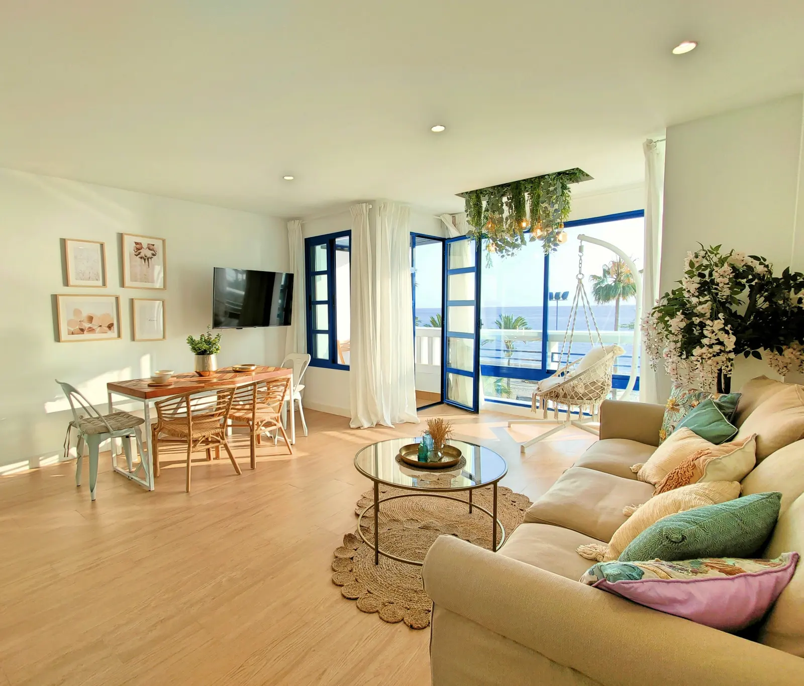 Salón comedor de paredes blancas y suelo de madera clara, con mesa de comedor y sillas de mimbre, sofá con cojines, mesa de centro de cristal, televisor en la pared y puertas vidriera de marco azul abiertas a una terraza con palmeras y vistas al mar.