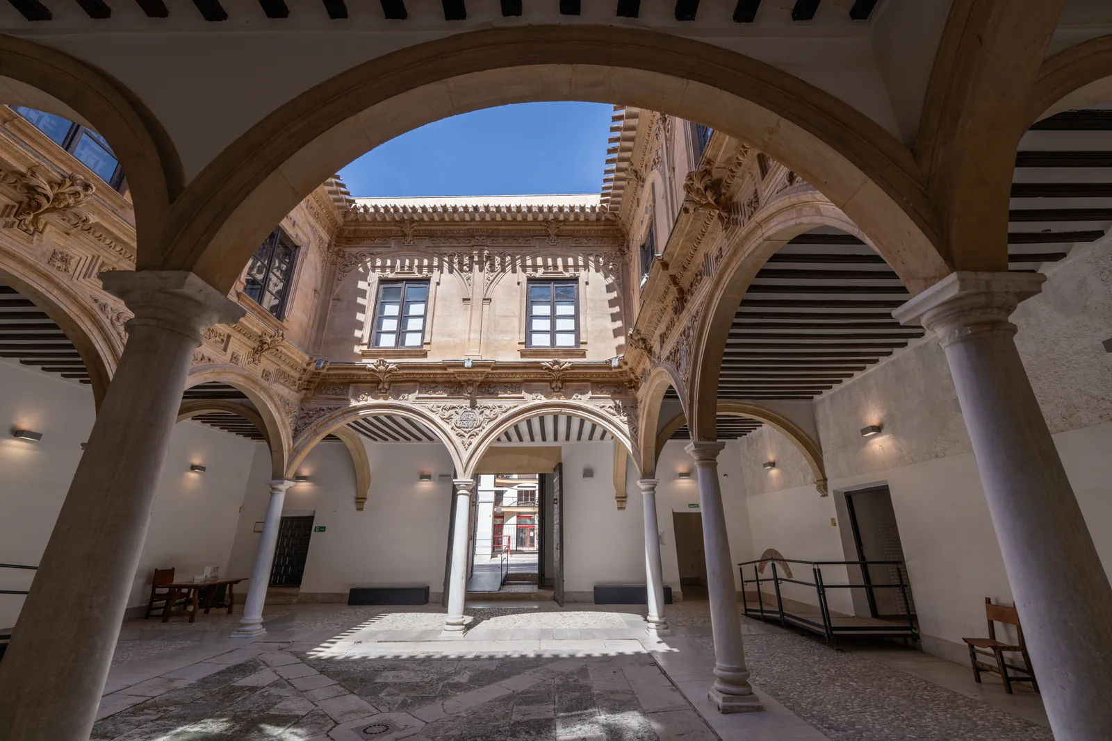 Patio interior porticado con arcos de medio punto sobre columnas de piedra, galería perimetral cubierta con vigas de madera, ventanas con rejería en el piso superior y suelo de losas de piedra; al fondo, una puerta abierta deja ver la calle.