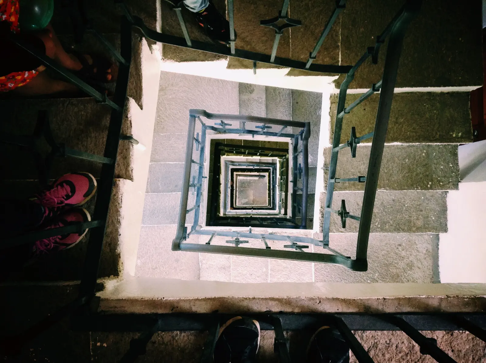 Vista en picado de un hueco de escalera de trazado cuadrado, con peldaños de piedra y zócalos claros, barandilla metálica oscura con motivos geométricos y varios rellanos superpuestos en profundidad, con pies de personas asomando por los bordes del encuadre.