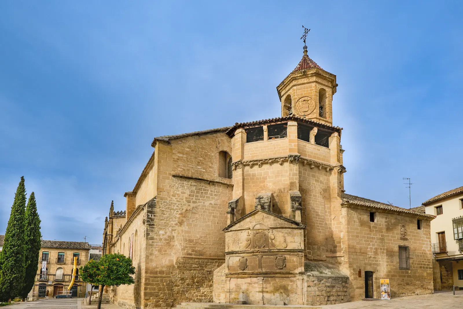 Edificio histórico de sillería con torre octogonal de chapitel y veleta, adosado a una plaza empedrada con dos cipreses, un naranjo y viviendas de ladrillo al fondo.
