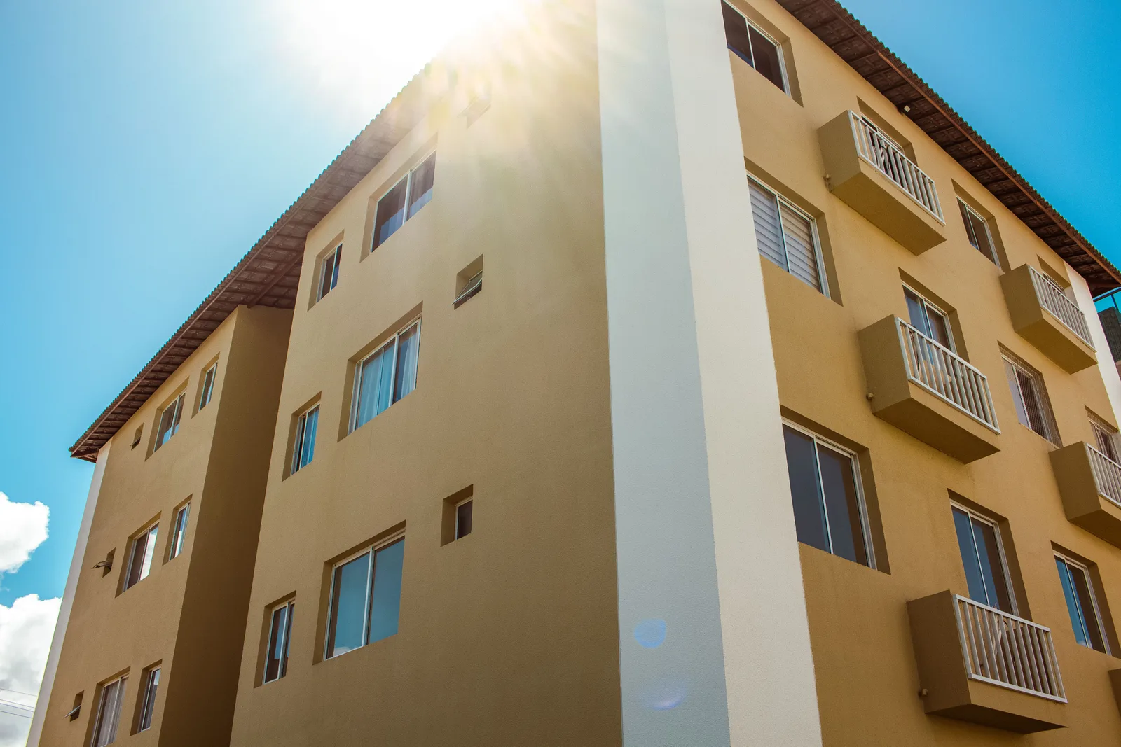 Fachada de un edificio residencial de color ocre con ventanas y balcones de barandilla blanca, vista en contrapicado bajo un cielo azul con sol de contraluz.