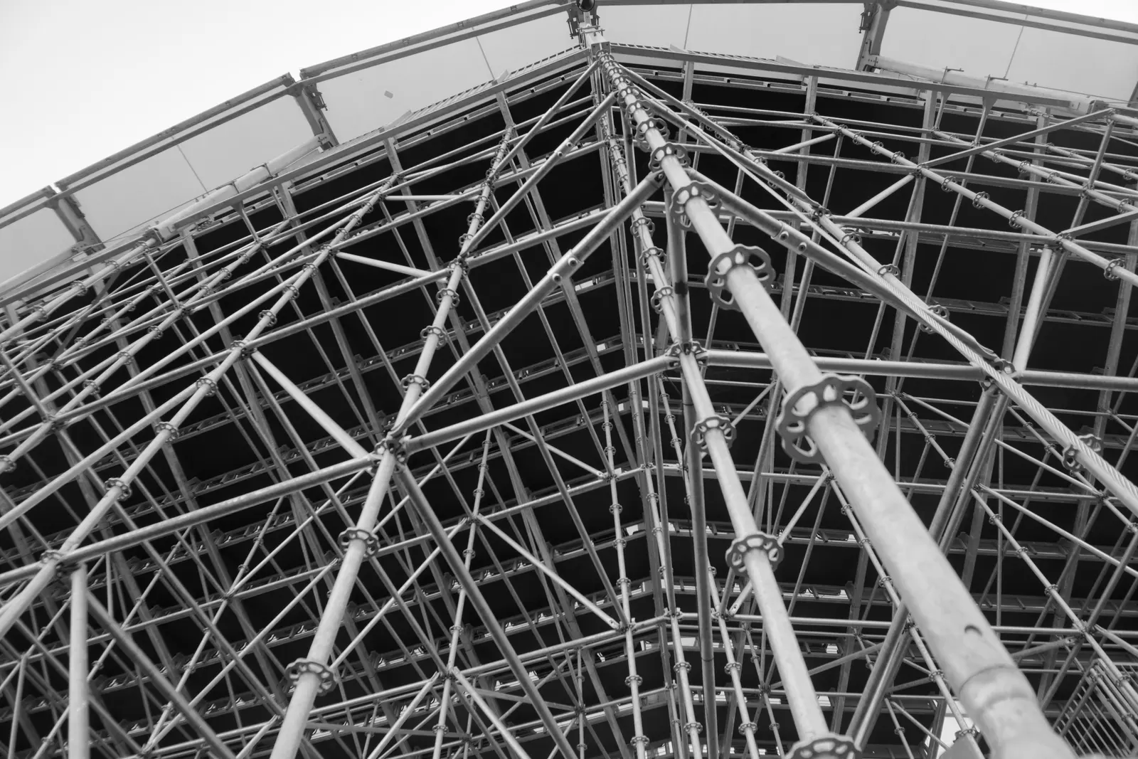 Estructura de andamio tubular metálico fotografiada en contrapicado, con barras verticales y horizontales ensambladas mediante abrazaderas de roseta, ante una fachada oscura y cielo claro al fondo, en blanco y negro.