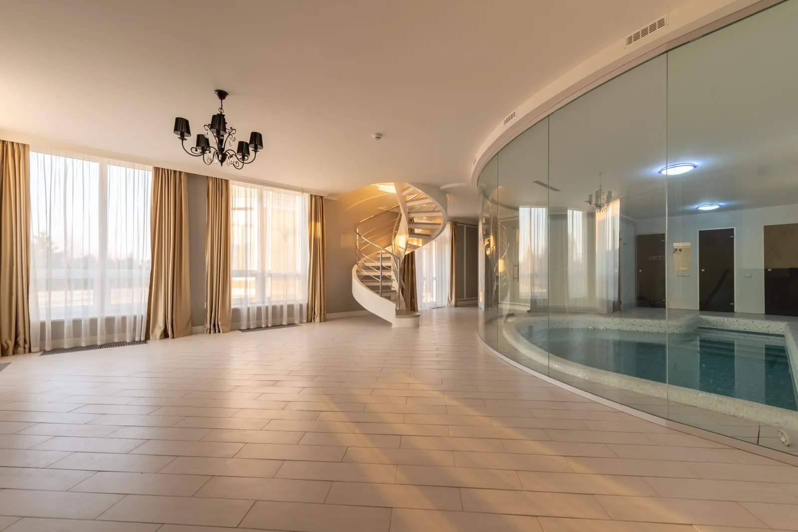 Salón diáfano de una vivienda con suelo de baldosa clara, grandes ventanales con cortinas, lámpara de araña, escalera helicoidal al fondo y una piscina cubierta separada por una mampara de cristal curvo.