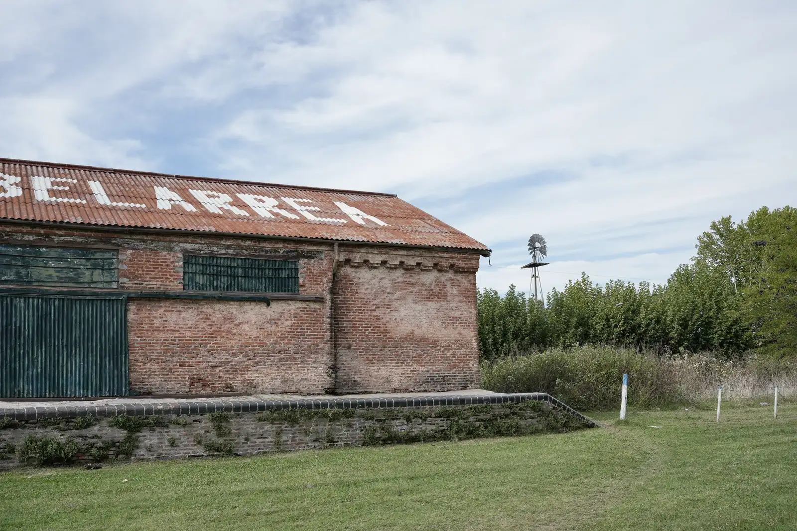 Vista exterior de un antiguo edificio de ladrillo visto con cubierta de teja roja, en la que se leen letras blancas pintadas sobre las tejas, con una gran puerta metálica verde y una ventana enrejada, sobre una plataforma de ladrillo con vegetación; al fondo, árboles, un molino de viento y cielo nub