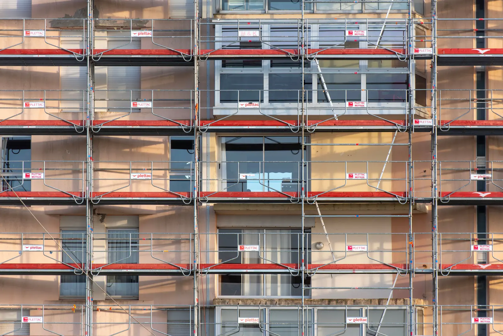 Fachada de un edificio residencial cubierta por un andamio tubular de varias alturas, con barandillas metálicas, plataformas de trabajo con bandas horizontales rojas y ventanas protegidas con barras.
