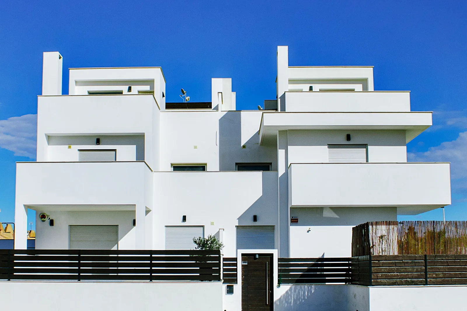 Fachada blanca de un edificio residencial de varias alturas con volúmenes escalonados, balcones salientes, persianas metálicas bajadas y algunos focos y detectores en el muro, ante un cielo azul con nubes y tras una valla de listones horizontales oscuros y una puerta de madera.