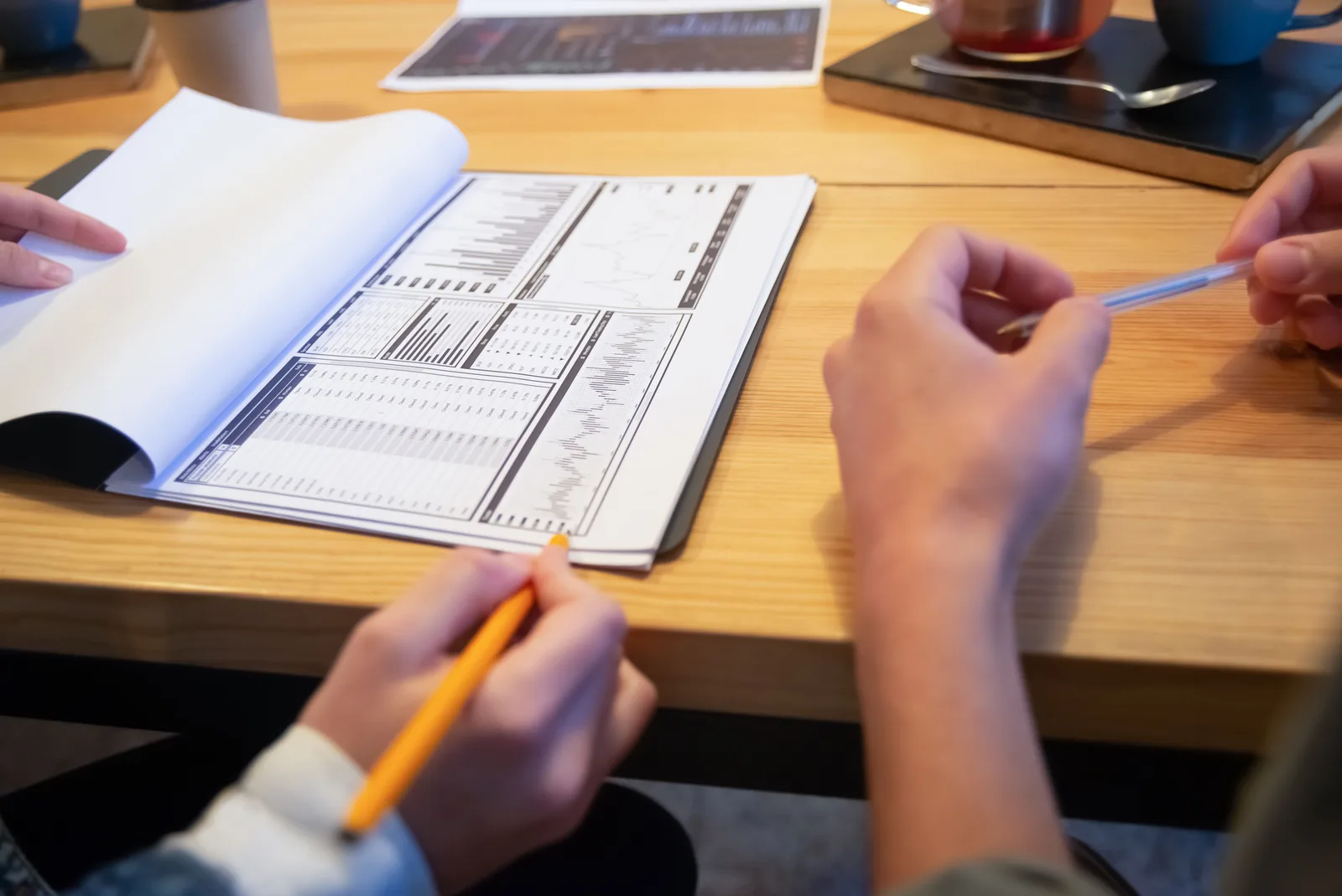 Manos sosteniendo un bolígrafo y un lápiz sobre una mesa de madera clara, con un cuaderno abierto y varios documentos impresos con tablas y gráficos, junto a una taza y una bandeja con utensilios al fondo.