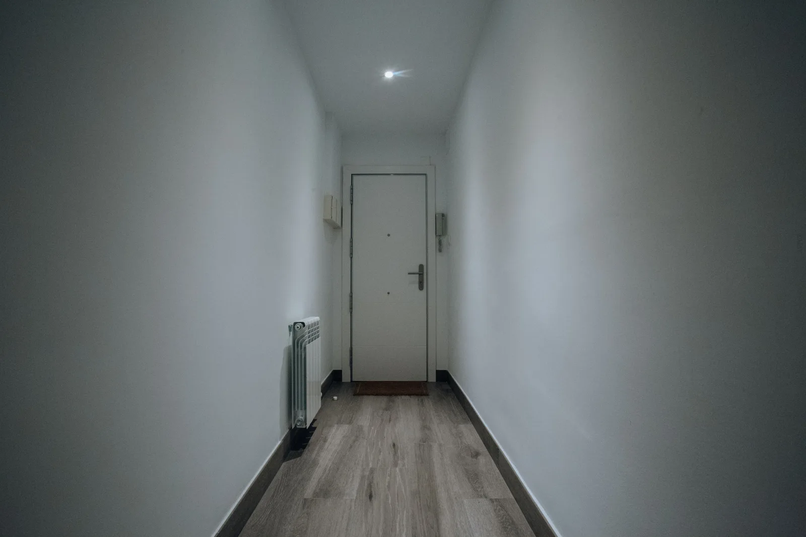 Pasillo estrecho de paredes blancas y suelo laminado imitación madera que termina en una puerta blanca cerrada, con un radiador blanco adosado a la pared izquierda y un pequeño punto de luz en el techo.